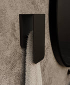 A white towel hanging on a matt black towel rack known as Aire mounted on a beige bathroom wall next to a round mirror with a black frame.