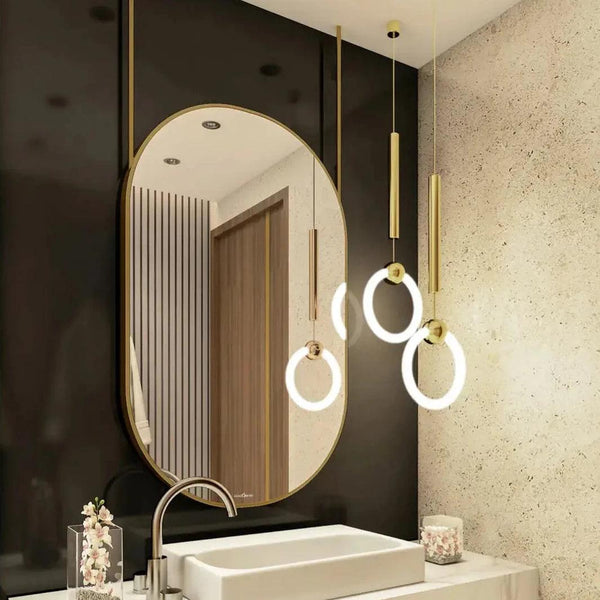 A close-up of a modern bathroom vanity set showcasing a mirror with a golden frame and is hanging on the roof through golden pipes. The mirror is mounted above a sleek, black countertop with a polished chrome faucet. 2 lights are also hanging from the roof with golden pipes and LED lights.