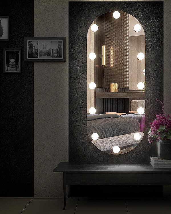 A lighted vanity mirror with a capsule shaped and has a frameless design. The mirror has bright LED lights around the frame and is positioned on a bedroom wall with a tabletop surface below it.