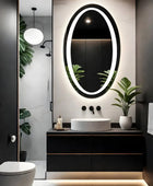 A simple bathroom featuring a rectangular white ceramic sink with a chrome faucet, a white toilet and a oval LED mirror with a frameless design mounted on the wall above the sink.