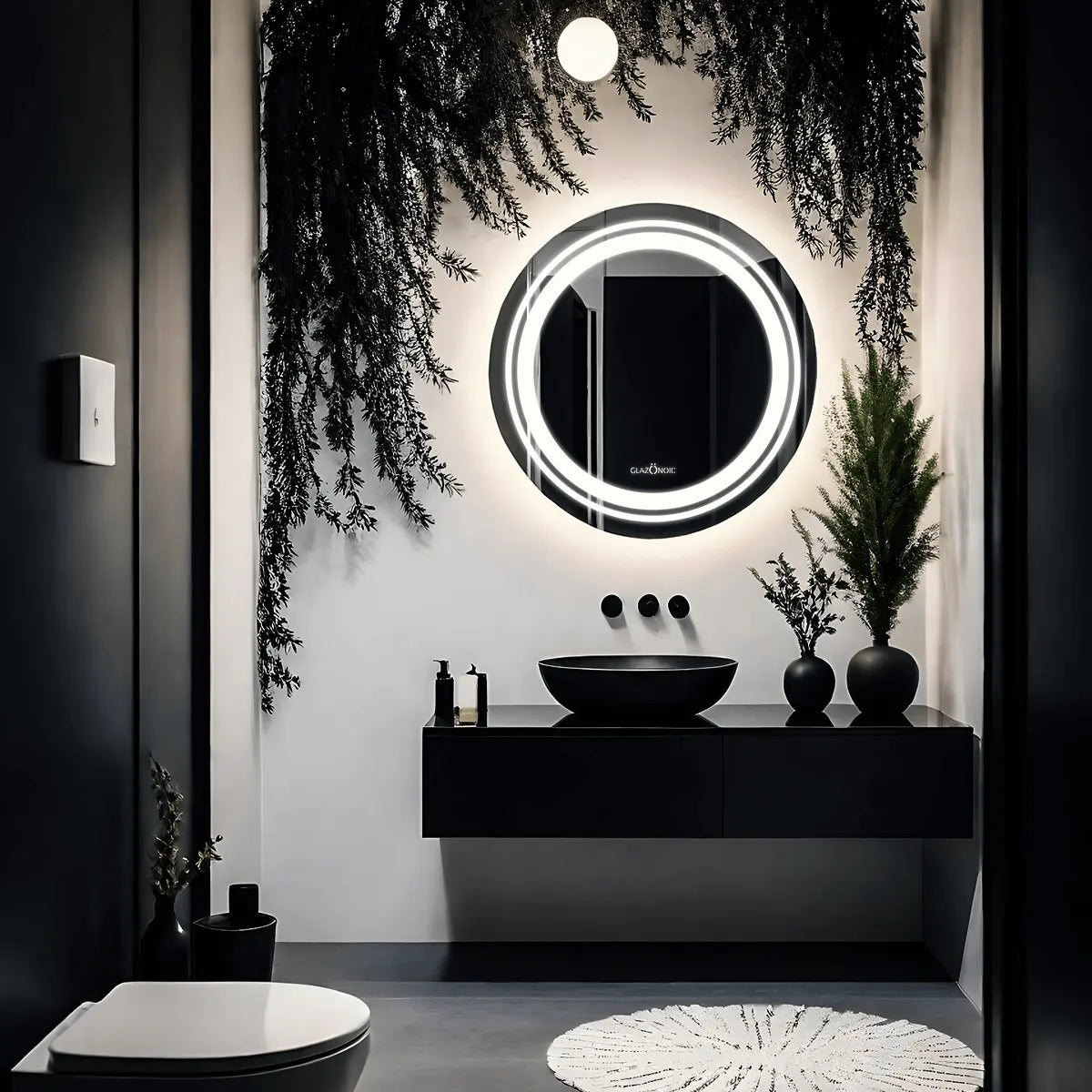 A close-up of a round bathroom LED mirror with a frameless design. The mirror is placed over the grey and black vanity which has a black ceramic sink and some plants with black pots. 