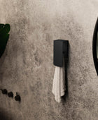 A white towel hanging on a matt black towel rack known as Aire mounted on a beige bathroom wall next to a round mirror with a black frame.