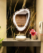 A close-up of a well-organized modern bathroom shelf featuring a white ceramic soap dispenser and two glass jars with chrome lids filled with cotton balls and cotton swabs, Along with a pebble shaped LED mirror with a nice and vibrant LED strip.