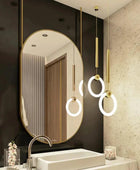 A close-up of a modern bathroom vanity set showcasing a mirror with a golden frame and is hanging on the roof through golden pipes. The mirror is mounted above a sleek, black countertop with a polished chrome faucet. 2 lights are also hanging from the roof with golden pipes and LED lights.