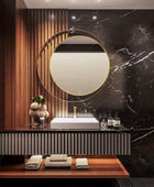 Modern and elegant bathroom vanity with a round mirror featuring a stainless steel gold colored frame and a sleek white marble sink countertop. With a plant and some perfumes kept on the counter top.