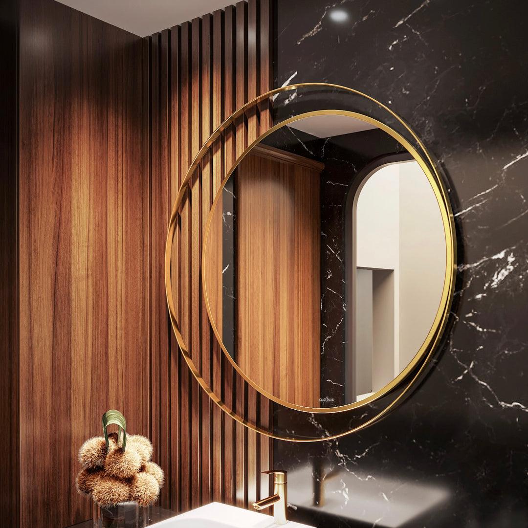 Modern and elegant bathroom vanity with a round mirror featuring a stainless steel gold colored frame and a sleek white marble sink countertop. With a plant and some perfumes kept on the counter top.